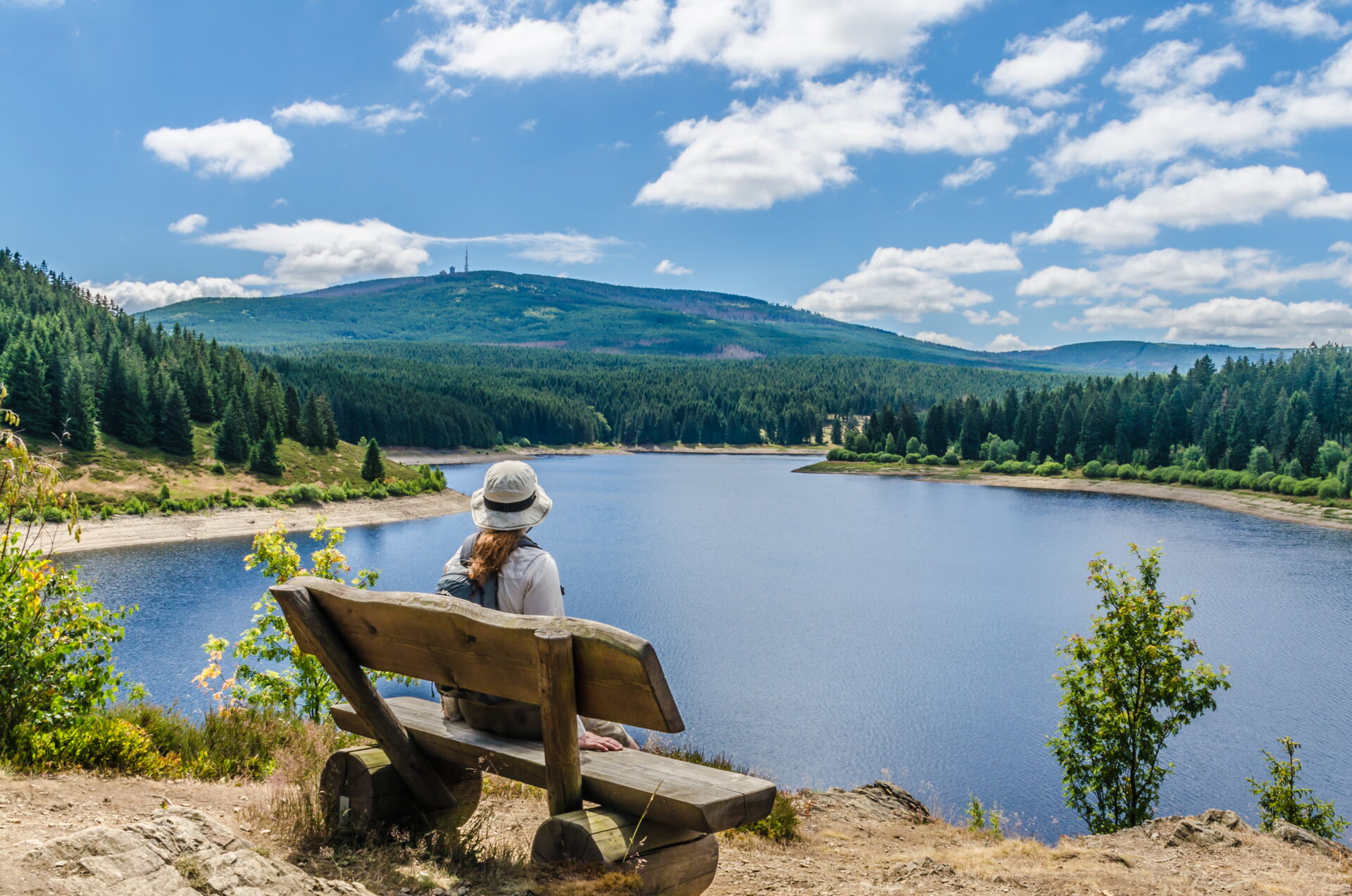 Eine Person mit Hut und Rucksack sitzt auf einer hölzernen Bank mit Blick auf einen ruhigen, blauen See, umgeben von grünen Wäldern und einem bewaldeten Hügel unter einem blauen Himmel mit weißen Wolken.