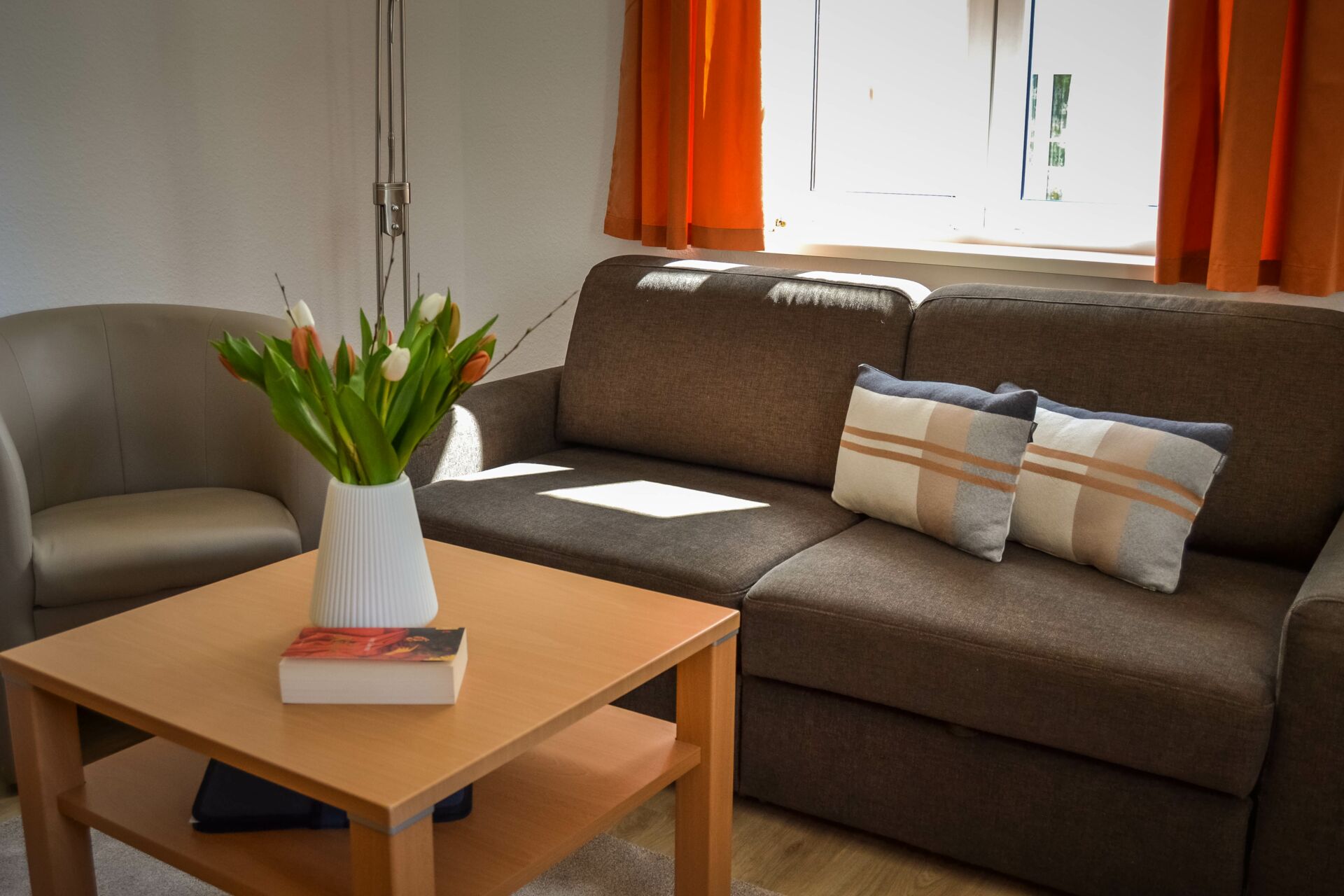 Ein gemütlicher Wohnraum mit einem braunen Sofa, zwei gemusterten Kissen, einem beigen Sessel und einem hellbraunen Couchtisch mit einer weißen Vase mit bunten Tulpen und einem Buch.