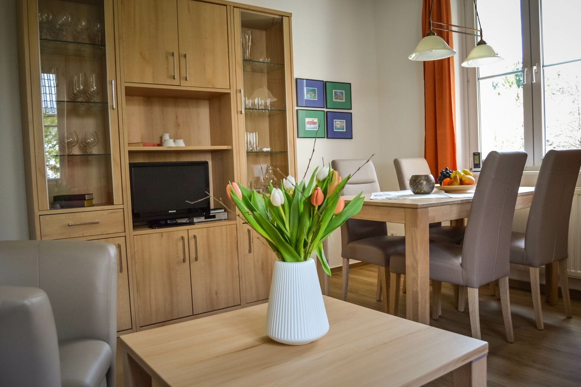 Ein hell eingerichteter Wohn- und Essbereich mit einem hellen Holz-Esstisch und vier Stühlen, einem passenden Wohnzimmerschrank mit Vitrinen und einem Fernseher, sowie einem Couchtisch mit einer weißen Vase mit bunten Tulpen.