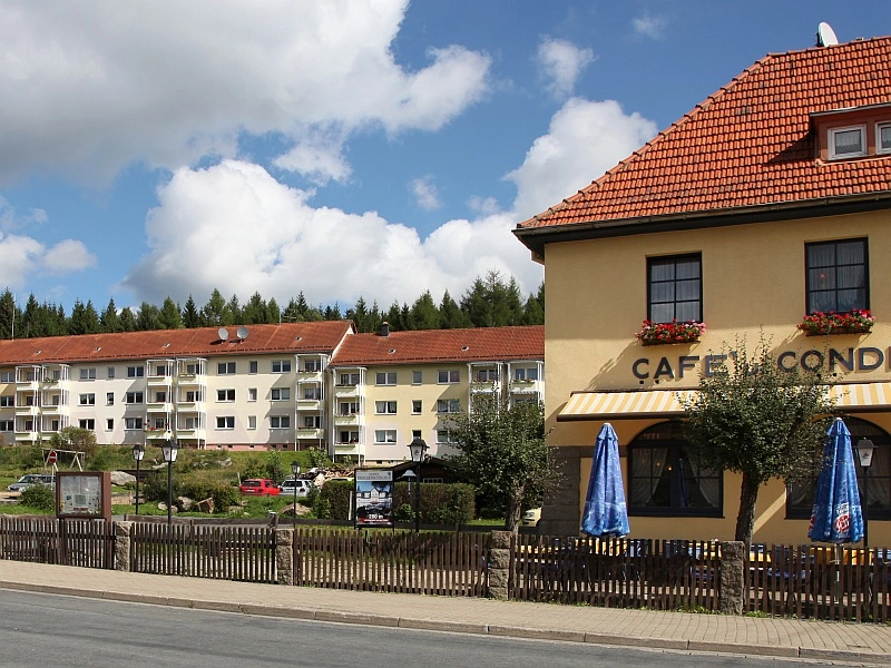 Eine Wohnanlage mit mehreren hellen Mehrfamilienhäusern und roten Ziegeldächern, eingebettet in eine grüne Umgebung mit Bäumen unter einem blauen Himmel mit weißen Wolken. Im Vordergrund ist ein gelbes Gebäude mit der Aufschrift "Cafe Konditorei" zu sehen