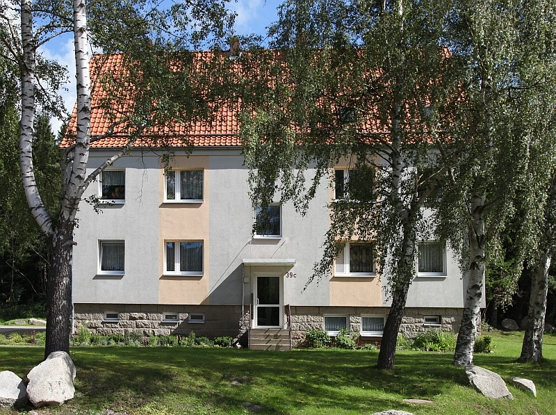 Ein zweistöckiges Wohnhaus mit einem roten Ziegeldach, einer hellgrauen Fassade und einer steinernen Basis. Mehrere Fenster sind sichtbar, und das Haus ist von Bäumen und einem grünen Rasen umgeben.
