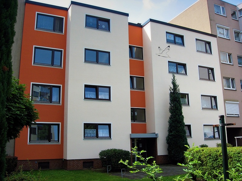 Ein modernes Mehrfamilienhaus mit einer Fassade in Weiß und Orange. Vor dem Haus befindet sich ein gepflegter Grünbereich mit Büschen und einem kleinen Baum.