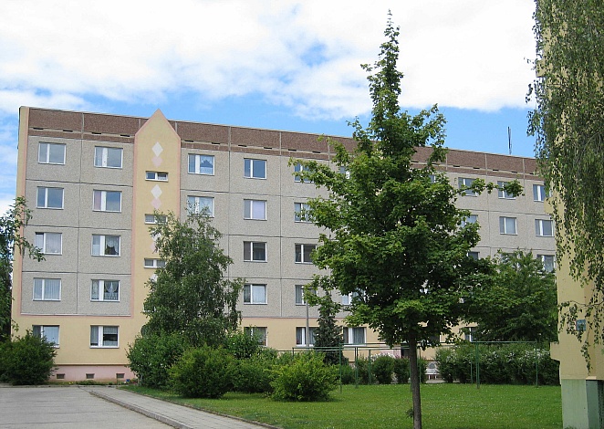 Ein Mehrfamilienhaus mit einer hellen Fassade und weißen Fenstern, umgeben von Grünflächen.